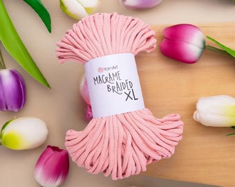Op de afbeelding: Een kluwen roze macramé gevlochten XL garen met een wit etiket met de tekst "Macrame Braided XL". Het garen is omgeven door kleurrijke tulpen met groene stelen en bladeren.