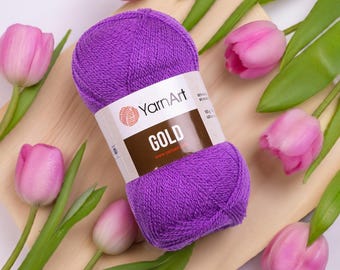 Op de afbeelding: Een bol paars YarnArt GOLD garen, 100g, ligt op een houten plank, omringd door roze tulpen en groene bladeren. Het garenlabel is zichtbaar, met de merknaam en productnaam.