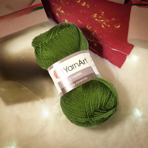 May include: A ball of green yarn with a label that reads "YarnArt ETAMIN". The yarn is a vibrant shade of green, and the label is white with brown accents. The yarn is likely used for knitting or crocheting projects.