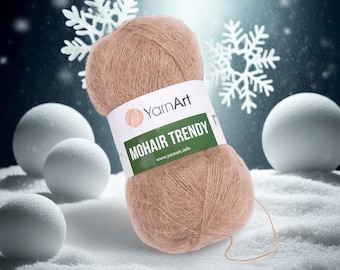 May include: A ball of light brown YarnArt Mohair Trendy yarn. The label is white with the brand logo and the text "MOHAIR TRENDY". The background features white snowballs and snowflakes, with a dark blue backdrop.
