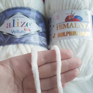 Puede incluir: Dos ovillos de hilo blanco, uno etiquetado como "alize Velluto" y el otro como "HIMALAYA DOLPHIN BABY". El hilo es suave y esponjoso, perfecto para tejer o hacer ganchillo.