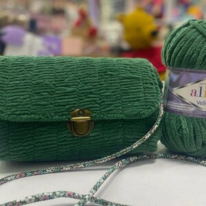 Puede incluir: Un bolso de mano de tela verde con un cierre dorado y una correa con estampado floral. El bolso est&aacute; al lado de una bola de hilo verde con la etiqueta "Alize Velluto" en ella.