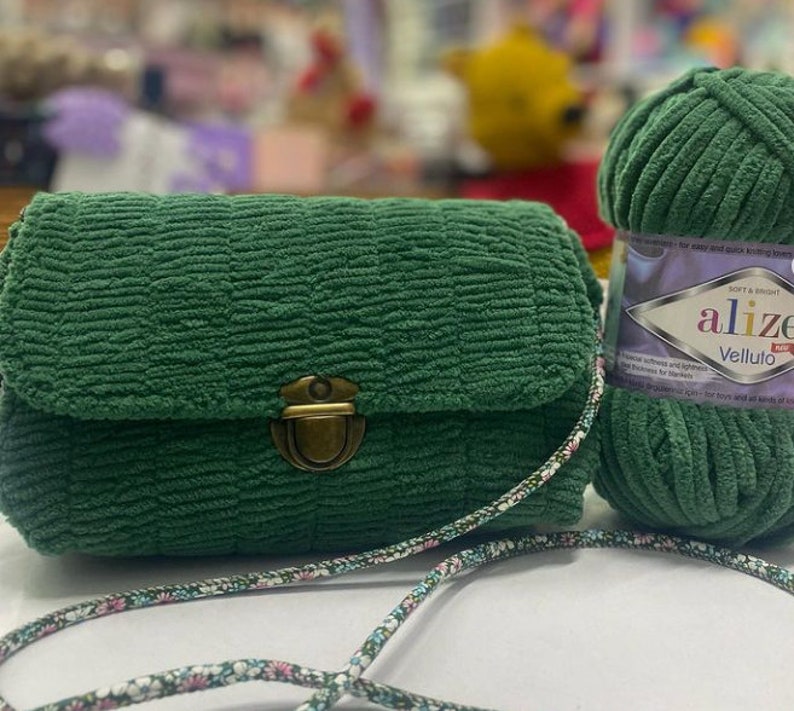 Puede incluir: Un bolso de mano de tela verde con un cierre dorado y una correa con estampado floral. El bolso est&aacute; al lado de una bola de hilo verde con la etiqueta "Alize Velluto" en ella.