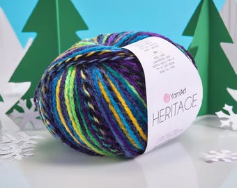 May include: A ball of YarnArt Heritage yarn in a mix of colours including blue, purple, yellow, and green. The yarn has a label with the brand name and product details. The background features green paper Christmas trees and snowflakes.