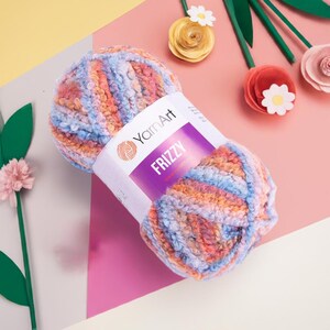 Op de afbeelding: Een bol veelkleurig garen met het label "Frizzy" van YamArt. Het garen is een mix van blauwe, oranje en roze tinten. De achtergrond is een mix van roze, geel en wit, met papieren bloemen.