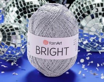 Op de afbeelding: Een zilveren bol garen met een wit etiket met de tekst "BRIGHT". Het garen is omgeven door discoballen. Het YarnArt-logo is ook zichtbaar op het etiket. De garenbol heeft een diameter van ongeveer 7,6 cm.