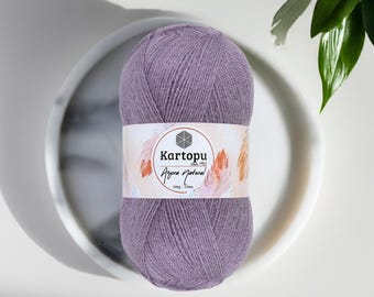 May include: A ball of lavender-colored yarn with a white label featuring the brand name "Kartopu" and the text "Angora Natural". The yarn is displayed on a white, marble-patterned plate, with green leaves in the background.