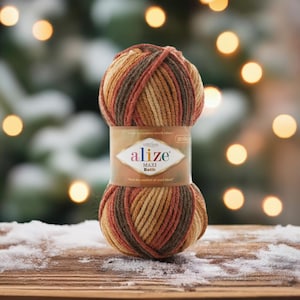 May include: A ball of Alize Maxi Batik yarn in shades of brown, orange, and tan. The yarn is wrapped with a label that reads "Alize Maxi Batik" and "Feel the comfort of wool blend". The background is blurred with bokeh lights.
