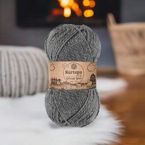 May include: A ball of grey Kartopu Melange Wool yarn, 100g/170m, with a brown label featuring a landscape illustration. The yarn is displayed on a white fur-like surface, with a blurred background of a fireplace and a woven basket.