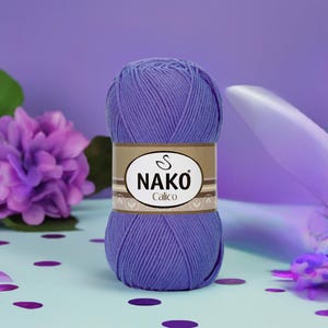 May include: A ball of lavender Nako Calico yarn. The yarn is wrapped with a brown label featuring the brand name and logo. The background includes a purple flower and a light blue surface with purple confetti.