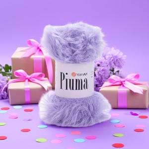 Op de afbeelding: Een bol zacht, lichtpaars YarnArt Piuma garen staat centraal, tegen een lavendelkleurige achtergrond. Twee ingepakte cadeaus met roze strikken en paarse bloemen flankeren het garen. Kleurrijke confetti stippelt het oppervlak af, wat een feestelijke scène creëert.