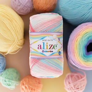 Può includere: Un gomitolo di filato Alize Bebe Batik dai colori pastello, tra cui rosa, blu, giallo e arancione. Il filato è avvolto con un'etichetta con il nome del marchio e una grafica di un bambino. Altre palle di filato di vari colori sono sparse intorno.