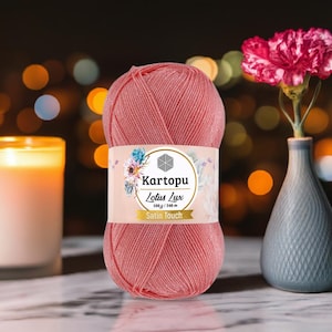 以下が含まれることがあります： 「Kartopu Lotus Lux Satin Touch」と書かれたラベルが付いたピンク色の糸玉。糸は100g / 340mです。ピンクの花が入った小さな花瓶と、火の灯ったろうそくが背景にあります。
