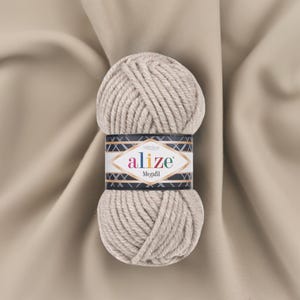 May include: A ball of Alize Megafil yarn in a neutral beige color. The yarn is thick and textured, with a label wrapped around the center. The label features the Alize logo and the word "Megafil". The background is a beige fabric.