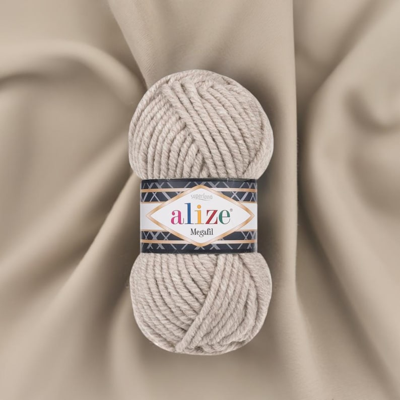 May include: A ball of Alize Megafil yarn in a neutral beige color. The yarn is thick and textured, with a label wrapped around the center. The label features the Alize logo and the word "Megafil". The background is a beige fabric.