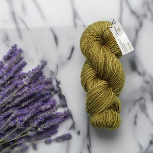 May include: A skein of olive green yarn with a tag that reads "Gazzal Worm". The yarn is displayed on a white marble surface next to a bundle of dried lavender.