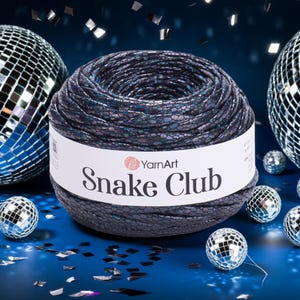 以下が含まれることがあります： ダークで虹色のYarnArt Snake Clubの糸玉。蛇の皮のような質感の模様があります。白いラベルが巻かれています。背景には、ディスコボールと紙吹雪が青い面に散りばめられています。