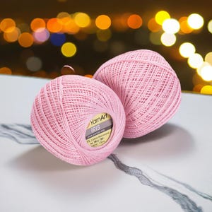 May include: Two pink yarn balls with the text "YarnArt IRIS" on the label. The yarn is a light pink color and appears to be cotton mercerized. The balls are on a white surface with a blurred background of yellow and orange lights.
