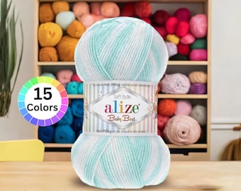 Puede incluir: Ovillo de lana Alize Baby Best en tonos azul claro y blanco. La etiqueta dice "Soft Bath" y "Anti-Pilling acrylic is used in this product." Un círculo cromático muestra "15 Colors." Estantes en el fondo están llenos de varios colores de hilo.