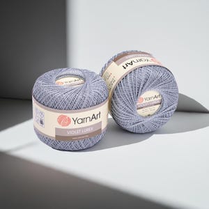May include: Two spools of YarnArt Violet Lurex yarn in a light purple colour. Each spool is wrapped with a label displaying the brand name and product details. The yarn is tightly wound, and the spools are positioned on a light-coloured surface.