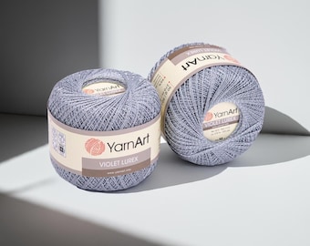 May include: Two spools of YarnArt Violet Lurex yarn in a light purple hue. Each spool is wrapped with a label displaying the brand name and product details. The yarn is tightly wound, and the spools are positioned on a light-colored surface.