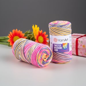 May include: Two spools of YarnArt Twisted Macrame 3mm yarn in a vibrant mix of pink, yellow, gray, and white. One spool is lying on its side, while the other stands upright. The label on the upright spool displays the brand name and product details. Decorative orange and yellow flowers are in the background.