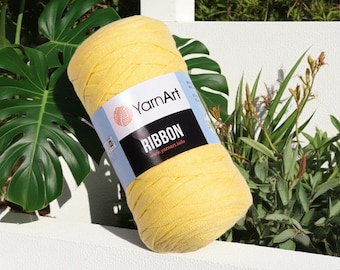 May include: A large, yellow skein of YarnArt Ribbon yarn. The label features the brand name and the word "RIBBON". The yarn is displayed outdoors with green foliage in the background. The yarn is a bright, solid color.