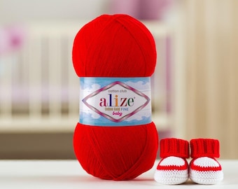 May include: A vibrant red ball of Alize Cotton Gold Fine Baby yarn with a label. Beside it are two small, handmade baby booties, red and white in color. The booties have a knitted cuff and a striped design.