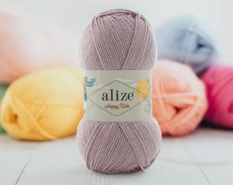 Puede incluir: Ovillo de lana Alize Happy Kids rosa empolvado. El hilo está envuelto con una etiqueta con el nombre y el logotipo de la marca. Se ven otros ovillos de lana en varios colores pastel en el fondo.