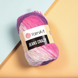 Puede incluir: Ovillo de lana YarnArt JEANS CRAZY en tonos rosa, morado y blanco. La etiqueta muestra el logotipo de YarnArt y el texto "JEANS CRAZY". La lana está bien enrollada, ideal para proyectos de tejido o ganchillo.