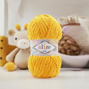 May include: A skein of bright yellow Alize Baby Softy Mega yarn. The yarn is wrapped with a label featuring the brand name and product details. A stuffed giraffe and other soft toys are in the background.