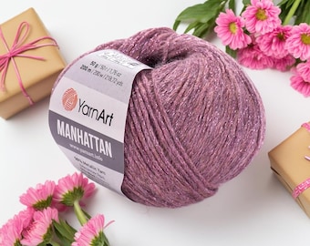May include: A ball of YarnArt Manhattan yarn in a mauve color with metallic threads. The label indicates 50g/1.76 oz and 200 m/218.72 yds. The image includes pink flowers and wrapped gifts.