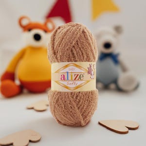 May include: A skein of Alize Softy baby yarn in a light brown color. The yarn is wrapped with a label that has the brand name and product details. In the background are two stuffed animal toys and wooden heart shapes.