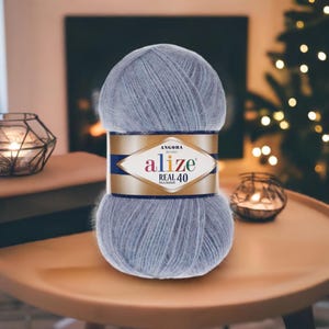 Peut inclure: Pelote de fil Alize Angora Real 40 bleu clair. L'étiquette affiche le nom de la marque et les détails du produit. Le fil est présenté sur une table en bois, avec un arrière-plan flou de cheminée et de sapin de Noël.