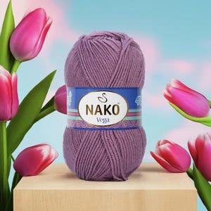 Puede incluir: Un ovillo de hilo NAKO Vega en un tono morado apagado, con una etiqueta azul y dorada. El hilo se exhibe sobre una superficie de madera, rodeado de tulipanes rosas con tallos y hojas verdes. El fondo es un suave degradado de azul y rosa.