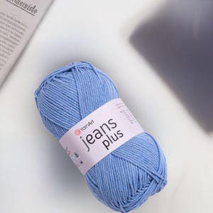 Op de afbeelding: Een bol lichtblauw garen met een wit etiket met de tekst "YarnArt jeans plus". Het garen is een zacht, getextureerd materiaal, ideaal voor brei- of haakprojecten. Het garen ligt op een wit oppervlak.