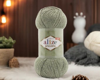 Puede incluir: Ovillo de lana Alize Alpaca Royal verde salvia. La lana está envuelta con una etiqueta que dice "Alize Alpaca Royal". La lana está sobre una superficie blanca y esponjosa con una piña en primer plano. Iluminación suave en el fondo.