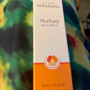 May include: A rectangular white and orange cardboard box for Avon Solutions Nurtura Serum. The front features the text AVON solutions Nurtura Serum/S&eacute;rum and a circular graphic with three colored shapes. The bottom indicates 30 ml / 1 fl. oz. U.S.