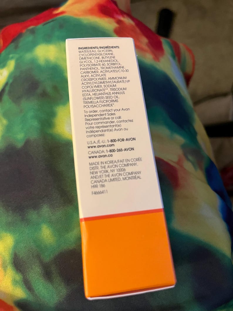 May include: A rectangular white product box with a bright orange base. The box features black printed text listing ingredients including water, glycerin, and sodium hyaluronate, along with contact information for Avon in the USA and Canada.