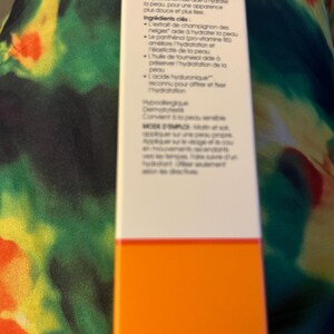 May include: A rectangular white skincare product box with black French text detailing ingredients like snow mushroom extract, panthenol, sunflower oil, and hyaluronic acid, featuring a solid orange horizontal band at the bottom.