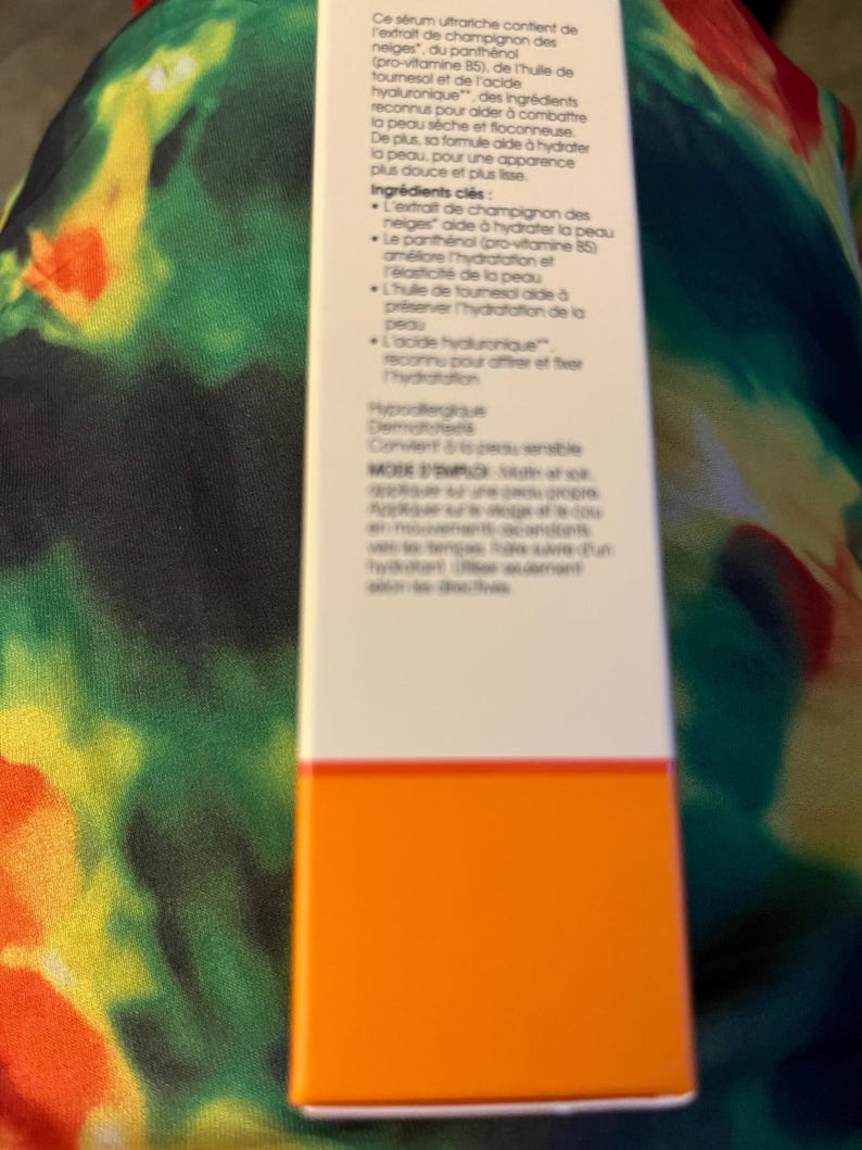 May include: A rectangular white skincare product box with black French text detailing ingredients like snow mushroom extract, panthenol, sunflower oil, and hyaluronic acid, featuring a solid orange horizontal band at the bottom.