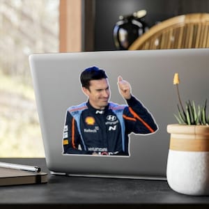 May include: A man in a blue and orange racing suit gives a thumbs up. He is wearing a helmet and has a Hyundai logo on his suit. The text "Hyundai Motorsport" is visible on the suit.