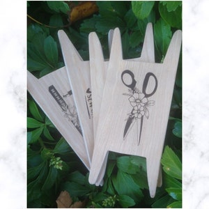 Puede incluir: Cuatro palitos de madera artesanales con diferentes diseños. El primer palito tiene una máquina de coser y un diseño de flores. El segundo palito tiene la palabra "Coser" con un diseño de flores. El tercer palito tiene un par de tijeras con un diseño de flores. El cuarto palito tiene una regla y un diseño de flores.