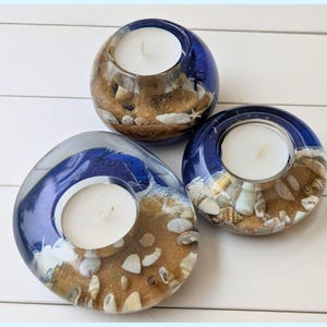 Pode incluir: Três castiçais decorativos com tema de praia. Cada um é feito de resina transparente com detalhes azuis, areia e conchas. Uma vela branca está dentro de cada castiçal, criando um ambiente costeiro.