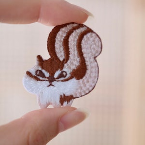 May include: An embroidered patch featuring a squirrel with a bushy tail. The squirrel is brown and white and has a serious expression.