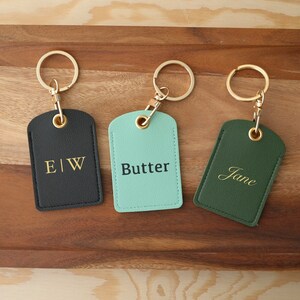 May include: Three leather keychains with gold rings and personalised text. The keychains are black, green and light blue. The text on the keychains reads "E|W", "Butter", and "Jane".