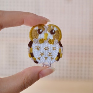 May include: A white embroidered owl applique with brown and yellow accents. The owl has large, round eyes with brown pupils.