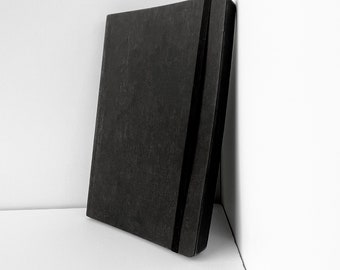 May include: A black notebook with a black elastic band closure. The notebook is lying on a white surface.