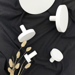 May include: White, round wooden knobs and a large, matching disc are arranged on a black fabric background. The knobs have a cylindrical stem and a flat, circular top. Dried wheat stalks add a natural element.