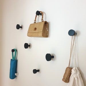 Può includere: Una collezione di ganci da parete neri con vari oggetti appesi. Una borsa beige con manico in bambù, un ombrello turchese piegato, un sacco bianco con coulisse e un rotolo di spago sono esposti. I ganci sono distanziati su una parete bianca.